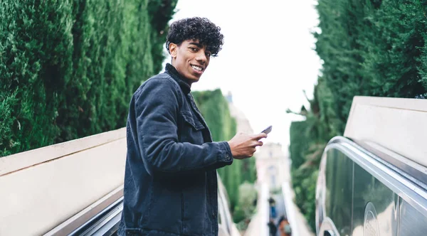 Gülümseyen kıvırcık kıvırcık saçlı siyah ceketli ve kot pantolonlu genç adam arka plandaki asansörde yeşil ağaçlarda kameraya bakıyor.