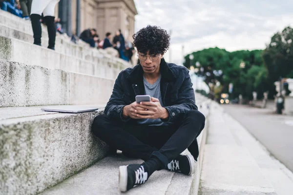 Młody Afroamerykanin Stylowej Odzieży Kręconymi Włosami Pomocą Smartfona Podczas Siedzenia — Zdjęcie stockowe