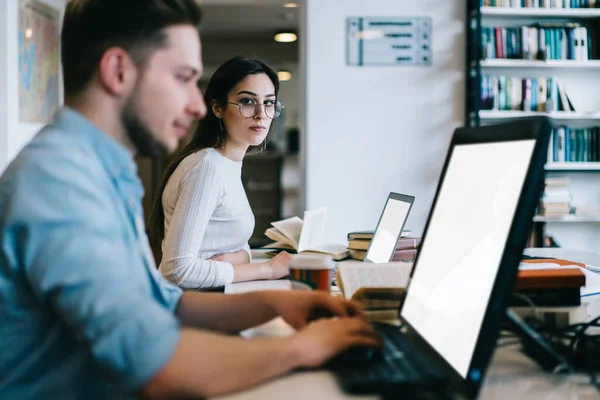 stock image Side view of diligent intelligent student typing on laptop while thoughtful woman in glasses looking for process at table in library