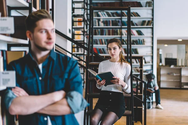 Entellektüel zeki, gündelik giysiler içindeki genç öğrenciler büyük şehir kütüphanesinde kitaplıkların karşısında duran kitapları seçip okuyorlar.