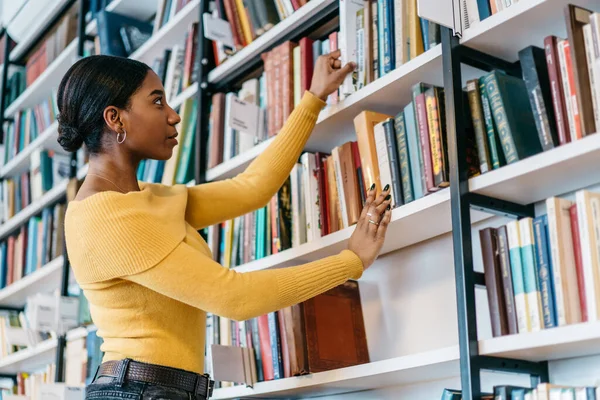 穿着休闲装的聪明的非洲裔美国女性在大学图书馆进行研究时从书架上取书的角度较低 — 图库照片