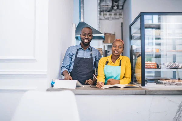 Happy successful confident African American owners of small confectionery shop standing at counter next to cake display fridge and looking at camera