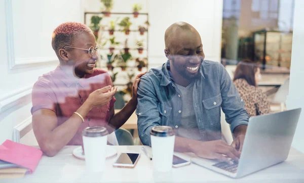 Through glass positive candid funny African American partners laughing on joke during cooperation about distance job with laptop in cafeteria