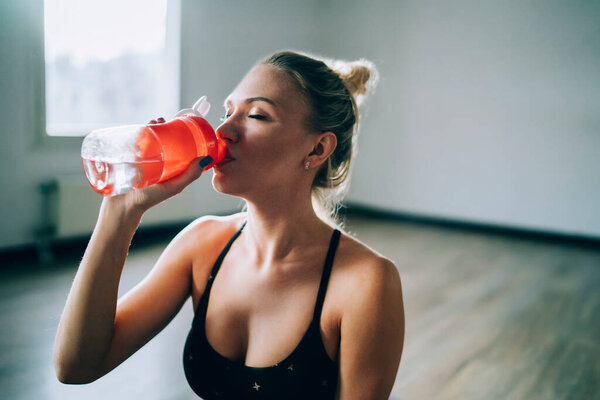 Shapely young woman in black sport bra relaxing and sipping water from bottle in studio in daylight with closed eyes