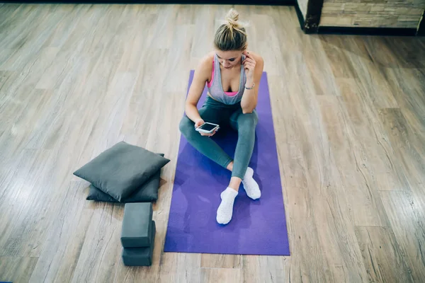 穿着运动服的金发女子坐在地板上的体操垫子上 看着智能手机的屏幕 — 图库照片