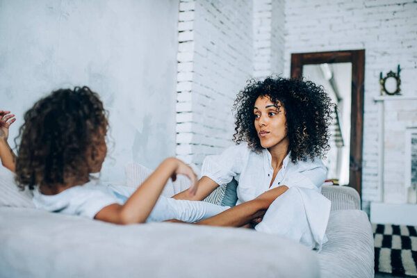 Playful curly haired mother in white clothes playing with daughter sitting on bed in studio with big mirror on wall in background