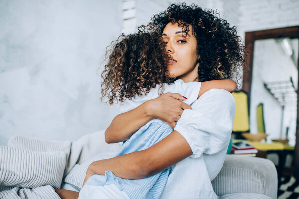 Beautiful tender woman sitting on cozy sofa and hugging curly haired little girl looking at camera in light studio 