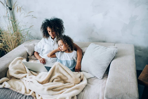 从上面看去 年轻而集中的母亲抱着可爱的小女儿 一边用笔记本电脑 一边躺在柔软的沙发上 毛毯铺在浅平的床上 — 图库照片