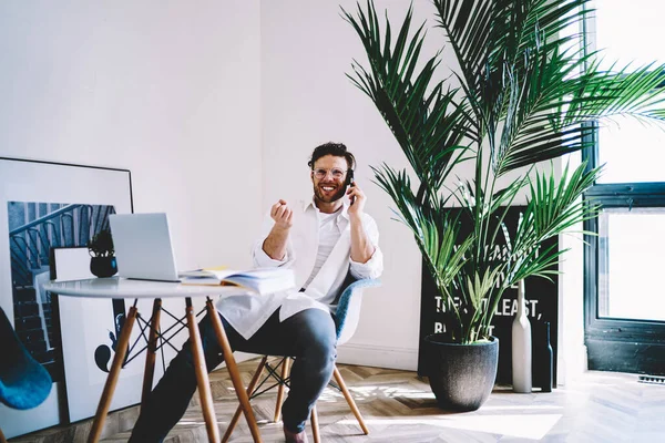 身穿眼镜 白衬衫和白衬衫的快乐而快乐的大胡子男性坐在创意工作室的椅子上打电话 — 图库照片