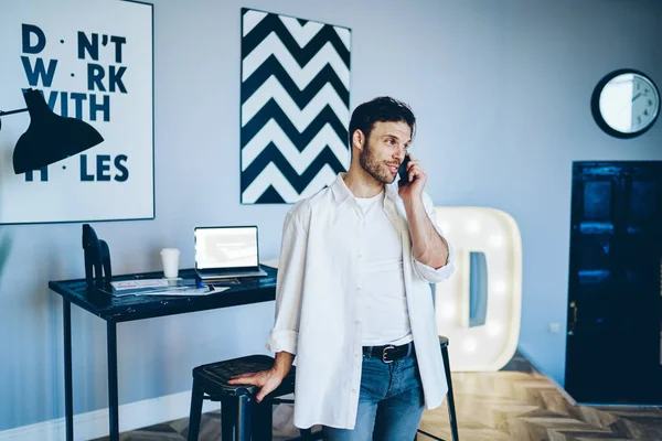 Happy hipster in white shirt and white t shirt making call on smartphone in creative workplace with black and white picture on wall