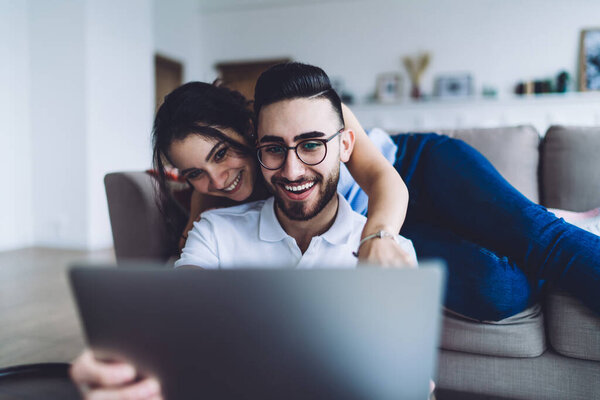 Positive smiling man and woman browsing laptop while resting on sofa by coffee table at modern living room in daylight