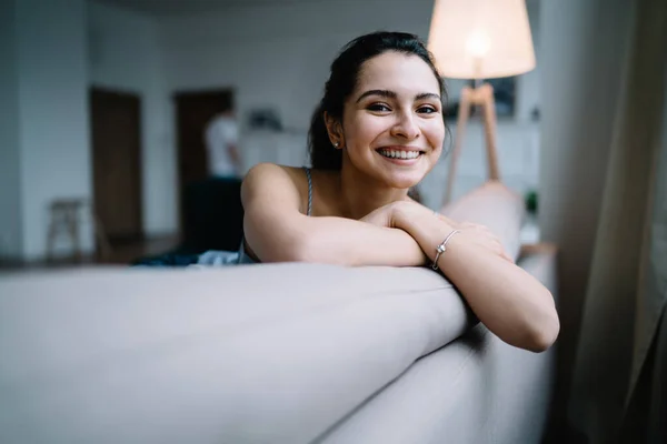 Cheerful woman with big smile leaning on sofa back with crossed arms and looking at camera on blurred background with apartment interior