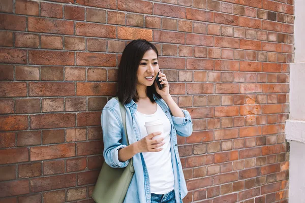 在电话交谈中 身穿牛仔裤衣服的自信的亚洲女人站在砖墙旁边 一边喝咖啡 一边用智能手机 — 图库照片