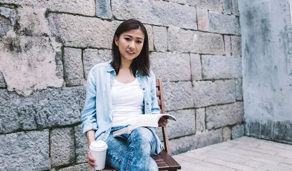 カジュアルな流行の気配り若いですアジアの女性とともにテイクアウトコーヒーカップ読書本ながら 日中の都市の通りに椅子に座って — ストック写真