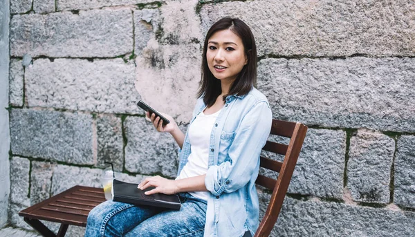 在中午时分 坐在砖墙旁边街道上的椅子上 随遇而安的年轻亚洲女人 一边拿着柠檬水 一边拿着外卖杯用智能手机发短信 — 图库照片