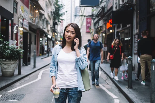 歩きながらスマートフォンで話すバッグを持ち カメラを見ているお店で路上でテイクアウトジュースを飲んでいるカジュアルなジーンズ姿でアジア系の大人の女性を笑顔 — ストック写真