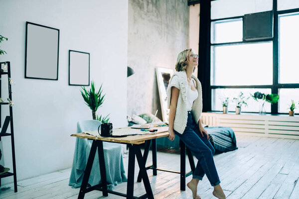 Side view of happy beautiful lady smiling while sitting on wooden table in middle of bright room and looking away