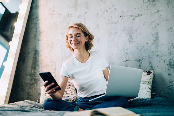 穿着休闲装 戴着眼镜 笑得漂亮的女性 一边用智能手机坐在床上 一边在客厅的笔记本电脑上浏览 — 图库照片