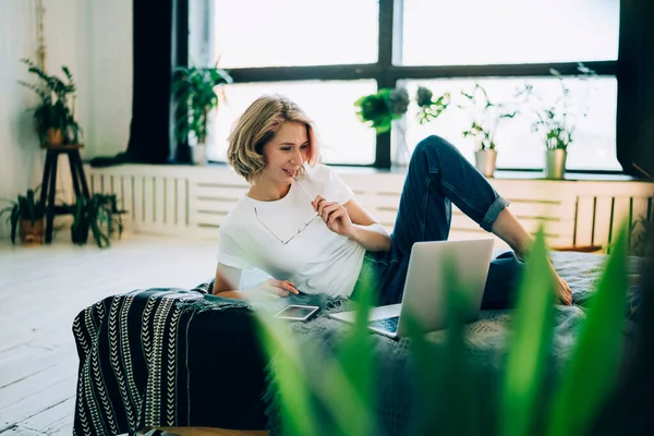Attractive hipster girl lying on comfortable bed and watching interesting motivation webinar on public website, beautiful woman enjoying resting time in home environment connceted to wifi internet
