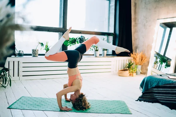 身穿运动服的体形瘦小的成年女性在绿色地毯上锻炼瑜伽 身体倒置 三脚架螺旋腿在家中的姿势 在阳光和植物的映衬下健康地生活 — 图库照片