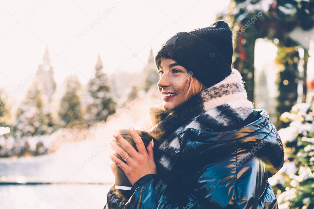 Side view of young merry woman in warm black clothes standing in cold snowy weather outdoors with paper cup with drink