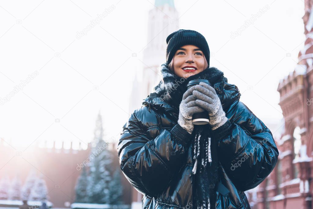 Optimistic delighted young woman in comfortable dark winter jacket standing near historical buildings with warm coffee in plastic cup on blurred background