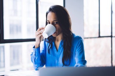 Düşünceli sakin Afro-Amerikan yetişkin kadın resmi kıyafetler içinde kahve içiyor ve açık modern apartmanın büyük penceresine odaklanıyor.