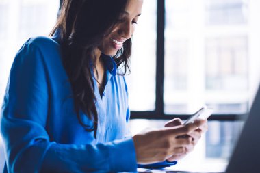 Afro-Amerikan zarif iş kadını heyecanlı bir şekilde cep telefonunda sörf yapıyor. Ofiste dizüstü bilgisayarının yanında oturuyor.