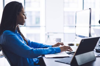 Masaüstünde büyük ekranlı rapor yazarken basılı diyagramlar ve dizüstü bilgisayarla çalışan konsantre genç siyahi kadının yan görünümü