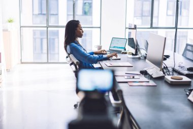 Siyahi iş kadınının iş hayatı hakkında tripodla film çekerken ofis masasında modern aletler kullanmasının yan görüntüsü.
