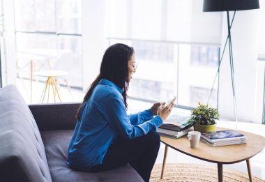 Gündelik kıyafetli genç siyahi bir kadının, modern evdeki kitap ve kupalarla koltukta oturup cep telefonuna bakması.