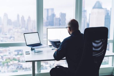Bilgisayarda çalışan gözlüklü kel adamın arka planda elinde bir yığın kağıt varken ofiste oturduğunu görüyorum.