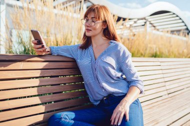 Çekici beyaz kadın açık havada akıllı telefon aracılığıyla mesajlar gönderiyor, güzel kadın blogcu parkta bankta oturan sosyal ağlarda yorumları okuyor.