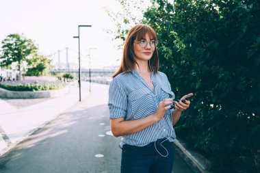 Güneşli bir günde, açık havada cep telefonuyla gezen göz elbiseli çekici kızıl saçlı bir kadın. Aksesuar, güzel bir yeniyetme kız, cep telefonundan sesli dinlemeyi seçiyor.