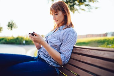 Bin yıllık düşünceli kızıl saçlı kadın parkta bankta oturan cep telefonuyla postaları kontrol etmeye odaklanmış. Kafkasyalı genç hippi kız, bankacılık ve online ödemeler için akıllı telefon uygulaması kullanıyor.