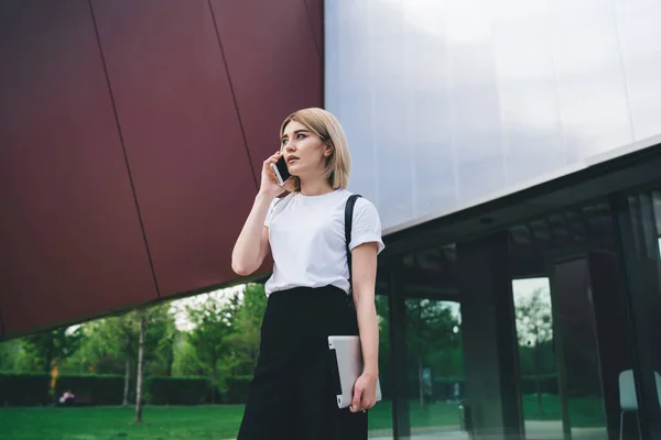 Pensive young modern woman in casual outfit and with black backpack holding tablet device talking on smartphone while walking against street building looking away