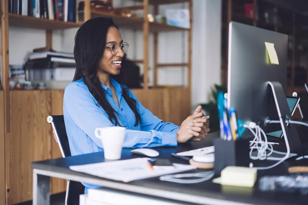 穿着蓝色正式衬衫 戴着眼镜 手放在桌子上看着电脑 微笑着坐在开放的办公室里工作的快乐的非洲裔美国女职员 — 图库照片