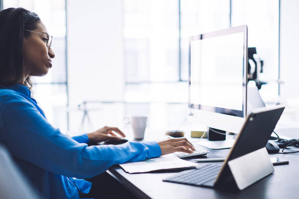 Side view of thoughtful African American female specialist looking attentively at reflective large computer screen in back lit while working with papers at table