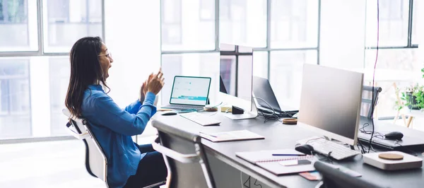 Side view of shocked African American adult female in formal clothes and glasses concentrating with open mouth on monitor while sitting and rising hands at table in light modern open space office