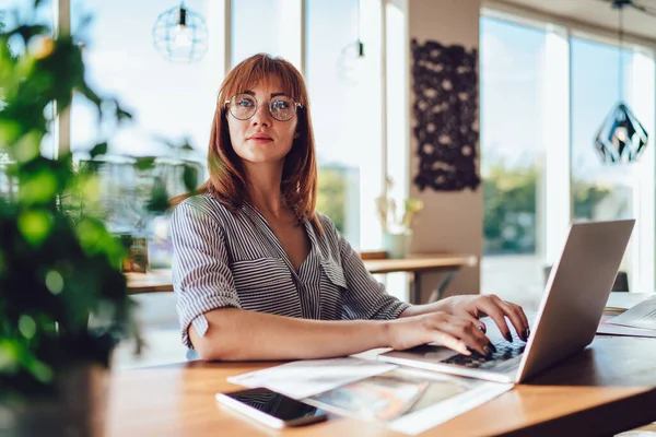 Attractive redhead woman in eyewear for vision protection working remotely typing publications sitting in cafe, pensive caucasian female freelancer look away browse on laptop computer during job