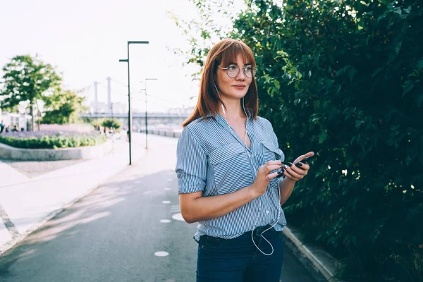 Atrakcyjna Ruda Kobieta Okularach Odtwarzających Zewnątrz Słoneczny Dzień Trzymając Telefon — Zdjęcie stockowe