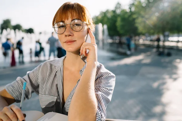 都市部の電話でノートに書く魅力的な白人の赤毛の女性の肖像 屋外で勉強するスマートフォンの会話を持つかなり千年の女性のヒップスター — ストック写真