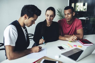 Modern iş yerinde masada otururken çeşitli erkek arkadaşlara proje detaylarını anlatan kalemle gülümseyen genç bir kadın.