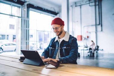 Neşeli erkek, gündelik giysiler içinde mutlu bir şekilde sırıtırken modern dijital aygıtlarla tavan arasında internetten bilgi topluyor.