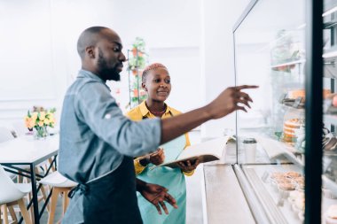 Not defteriyle sohbet eden ve kafeteryadaki tatlılara işaret eden Afro-Amerikalı genç, gülümseyen, neşeli meslektaşlar.