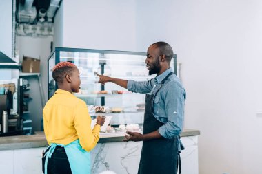 Önlüklü, not defterine not alan, birbiriyle sohbet eden ve kafeteryadaki tezgahı işaret eden Afro-Amerikalı genç, modern meslektaşlar.