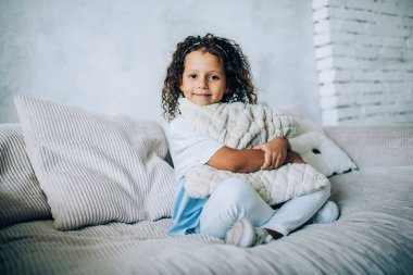 Gülümseyen, kıvırcık, tatlı bir çocuk. Yumuşak yastığını kucaklarken rahat bir şekilde rahat koltukta oturup kameraya bakıyor.