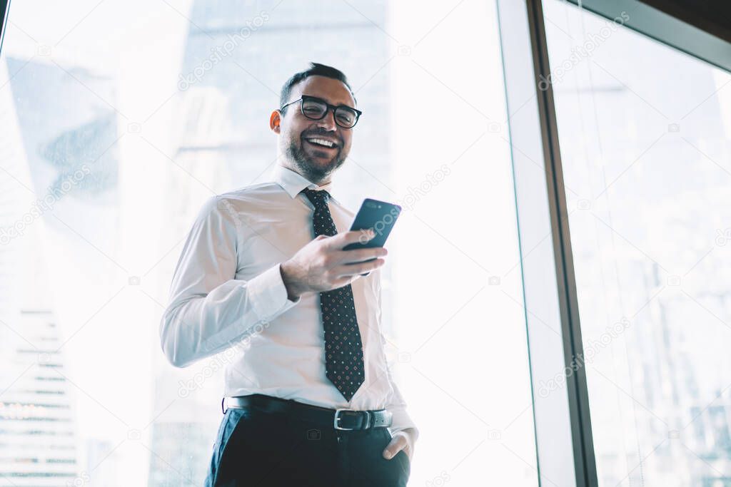 Half length portrait of cheerful male entrepreneur in formal clothing and optical spectacles for provide eyes protection smiling at camera during time for digital marketing via cellphone technology