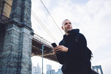 Aşağıdan, kısa saçlı, kıllı, siyah spor giysili, sırt çantalı, akıllı telefonlu genç bir adam. Şehir arka planında, devasa bir köprünün yanında yazılar yazıyor ve başka yöne bakıyor.