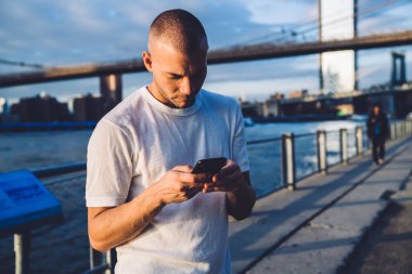 Genç yakışıklı, kısa saçlı, beyaz tişörtlü adam bulutlu bir sabahta köprünün yanındaki nehir kenarındaki kaldırımda duruyor ve akıllı telefondan mesaj atıyor.